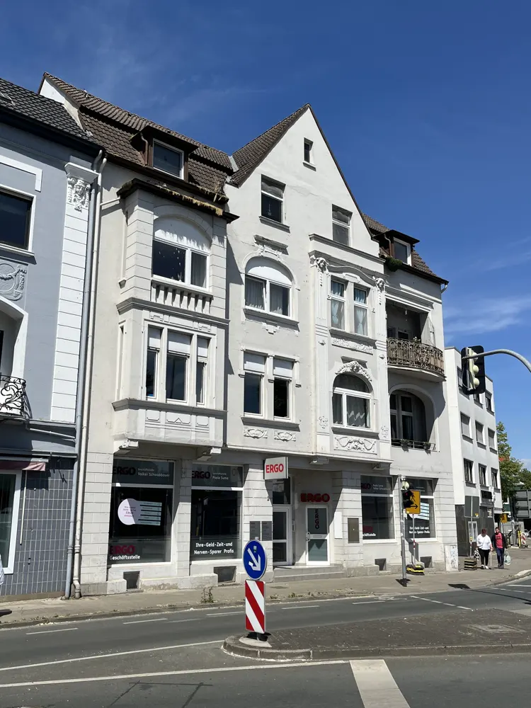 Three-story white building with an ERGO insurance office on the ground floor, street view, blue sky.