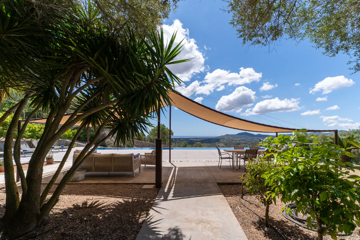 Outdoor patio with a beige sunshade, table, and seating overlooking a pool and distant hills under a blue, cloudy sky.