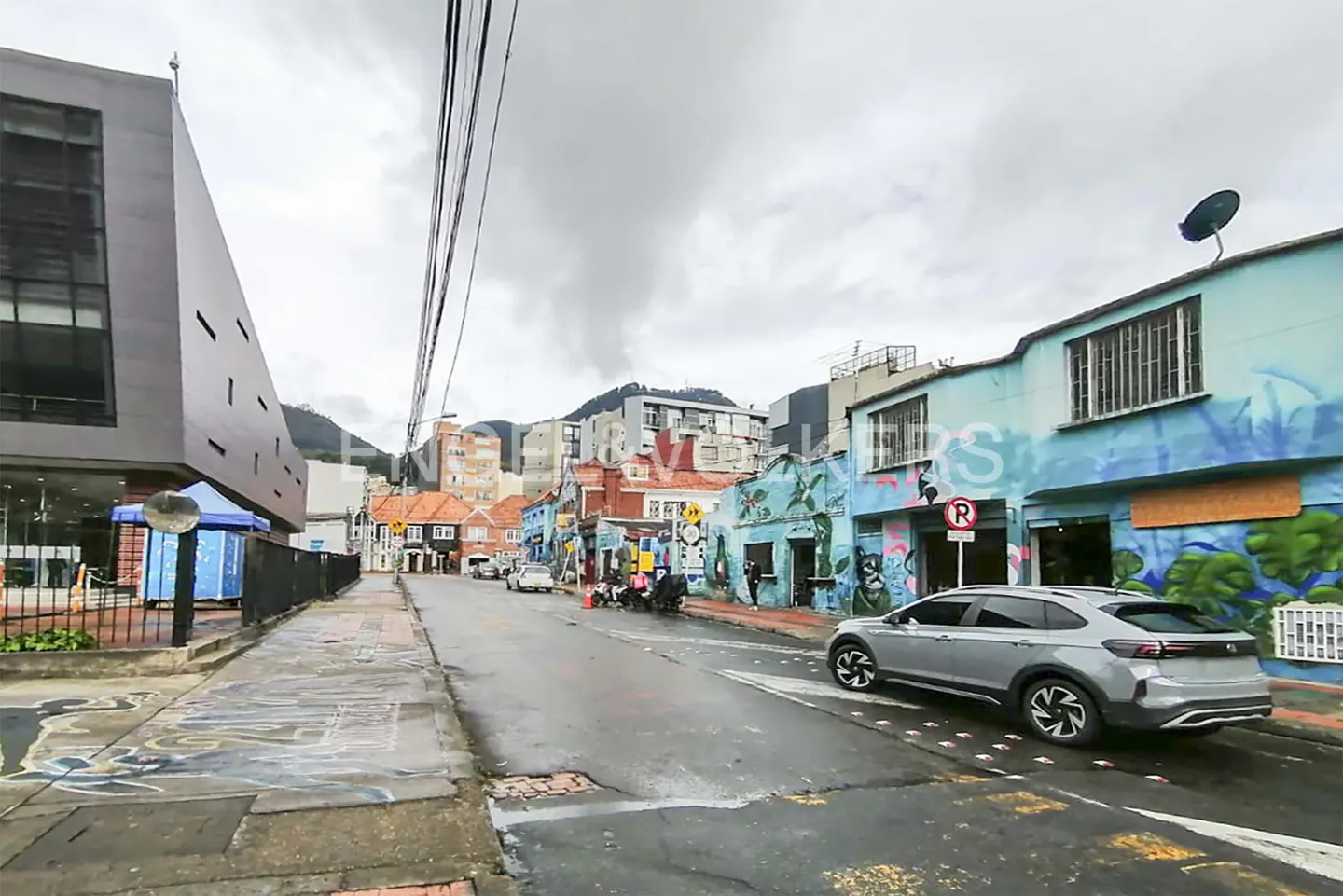 Street view of a gray car on a wet road, with colorful buildings and mountains in the background.