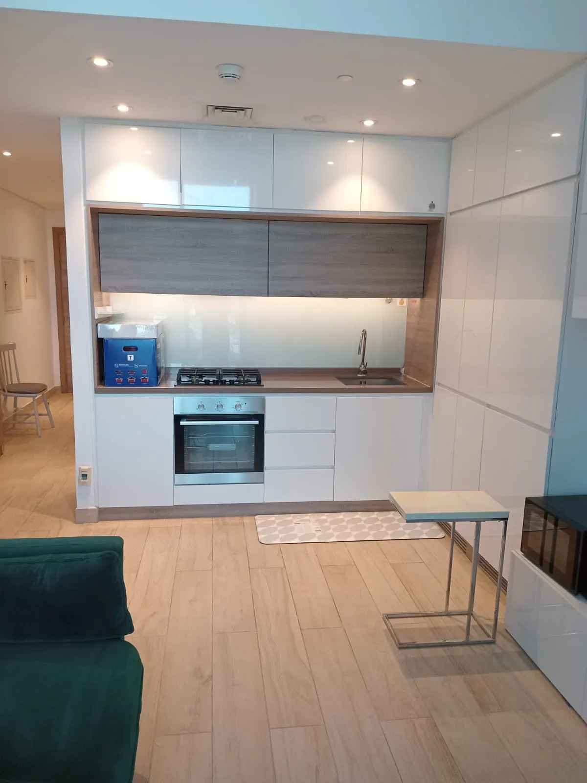 A modern kitchen with white cabinets, stainless steel appliances, and light wood floors. A green sofa is in the foreground.