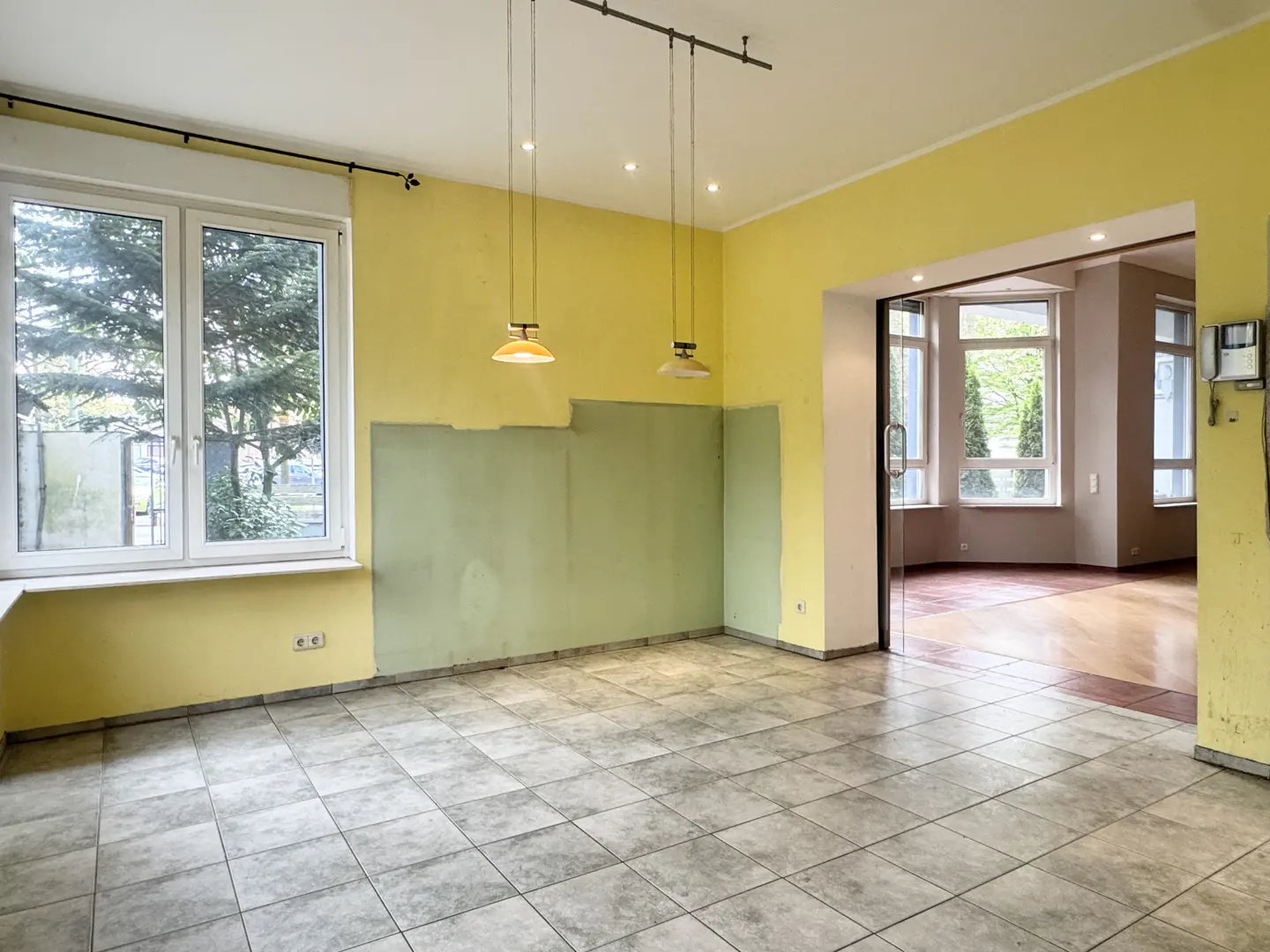 An empty room with yellow walls, a green accent wall, and gray tile flooring. Two pendant lights hang from the ceiling. A window and doorway are visible.