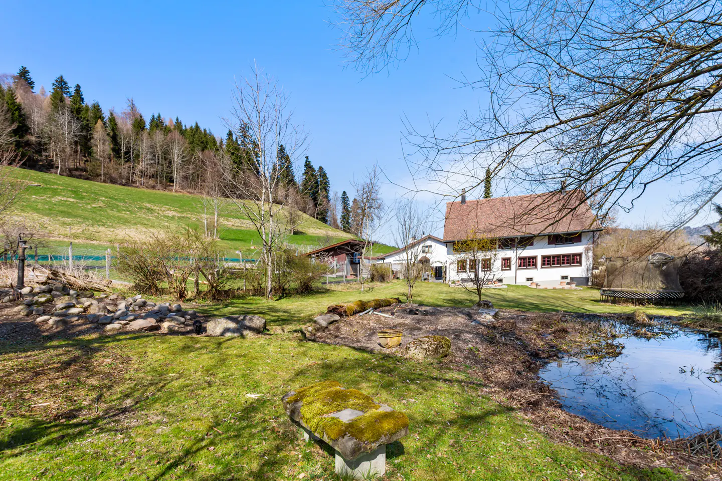 A white house with a red roof sits near a pond and green hill under a blue sky.
