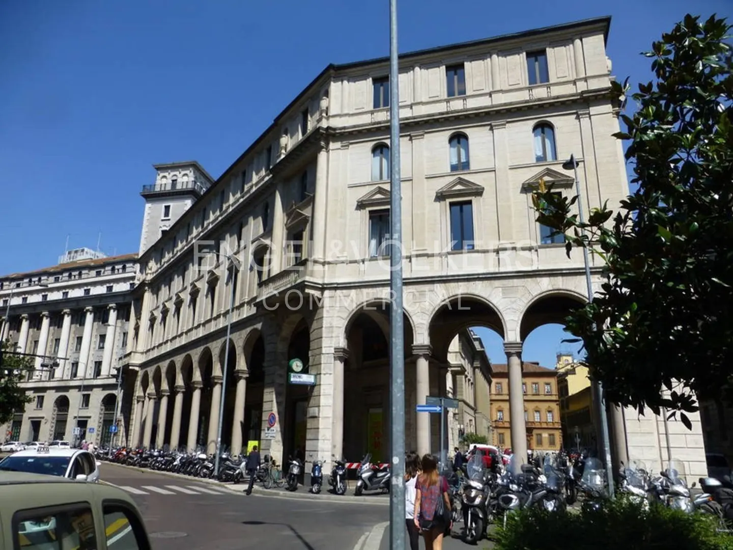 Street view of a long, light-stone building with arched walkways, parked motorcycles, and pedestrians on a sunny day.