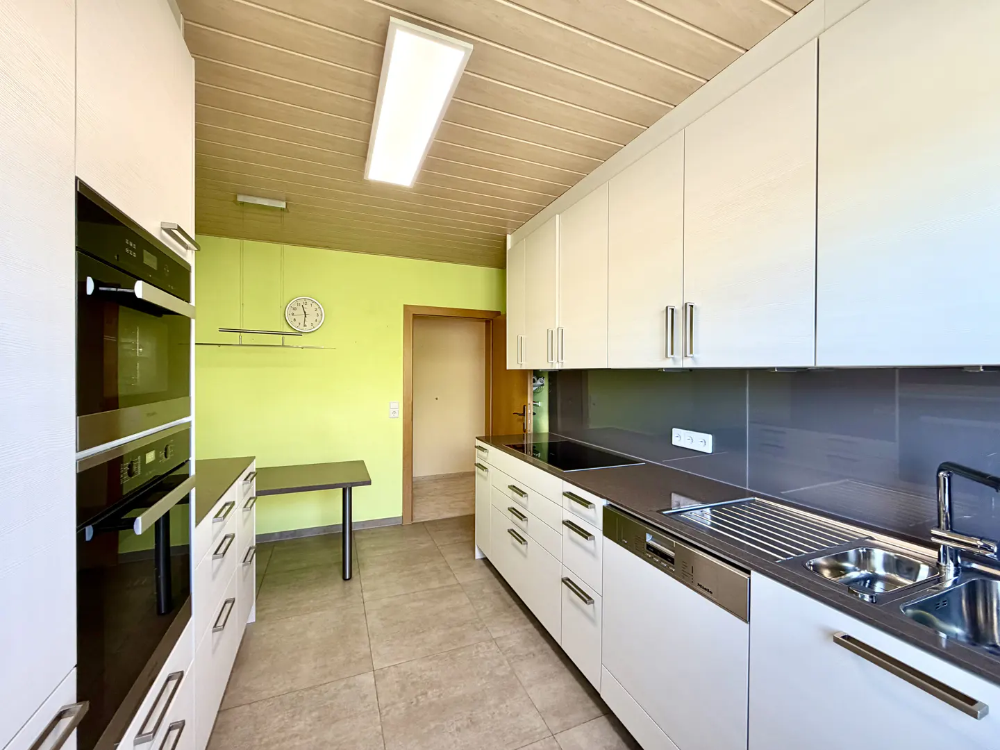 Bright kitchen with white cabinets, stainless steel appliances, and a gray countertop. A light green wall features a clock and a small table.