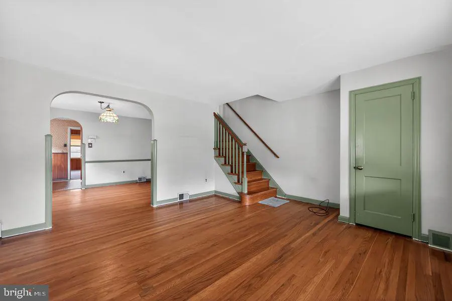A living room with hardwood floors, white walls, a green door, and a staircase. An arched doorway leads to another room.