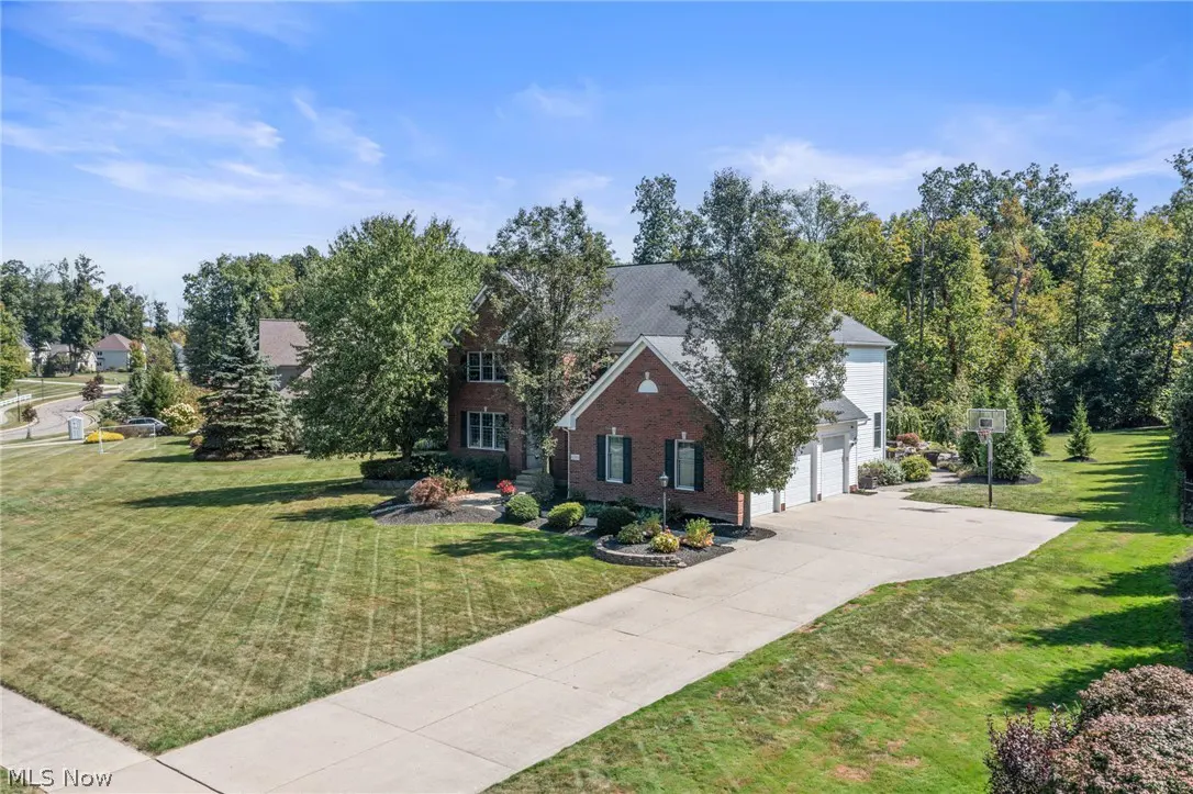 Aerial view of a two-story brick house with a gray roof, green shutters, and a long driveway surrounded by green lawns and trees.