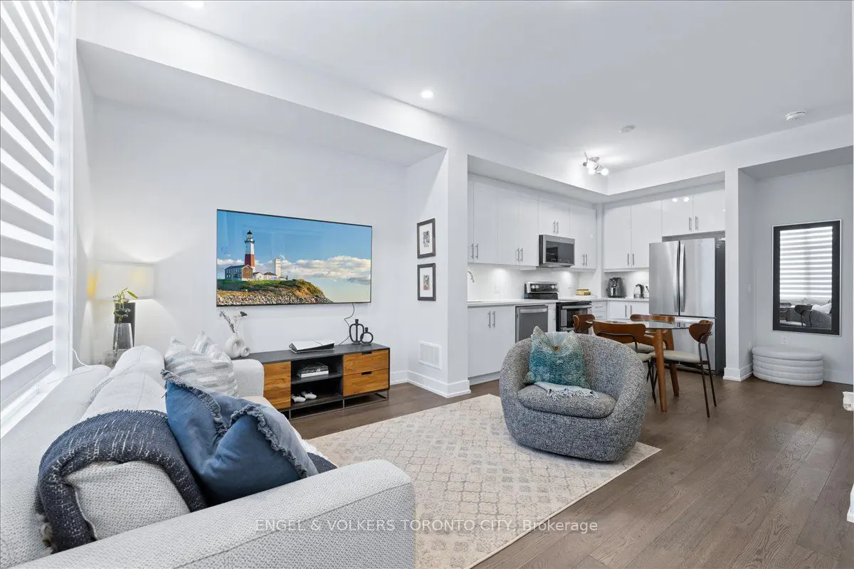 Bright living room with white walls, wood floors, and open kitchen. A TV displays a lighthouse scene. A gray sofa and chair offer seating.