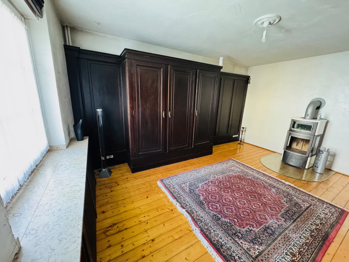 A room with wood floors, a large dark wood wardrobe, a red patterned rug, and a modern wood-burning stove.