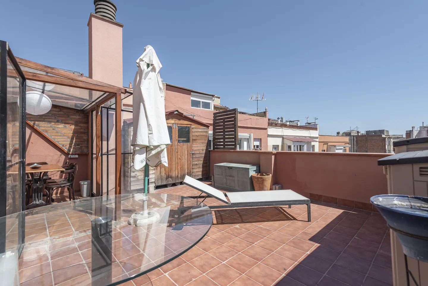 Rooftop patio with terracotta tiles, lounge chair, and glass table. A covered dining area and folded white umbrella are also visible.
