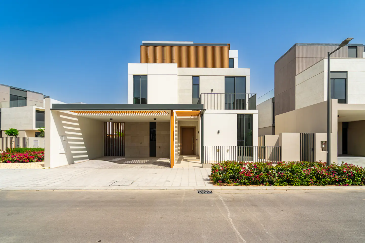 Modern two-story house with a carport, white walls, dark windows, and a wood-slat accent on the top floor. Red flowers in front.