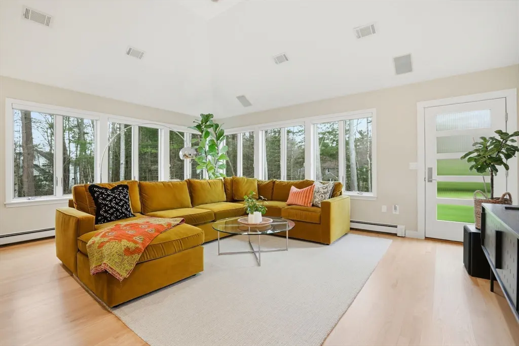 Bright living room with a large mustard-colored sectional sofa, a glass coffee table, and many windows overlooking a wooded area.