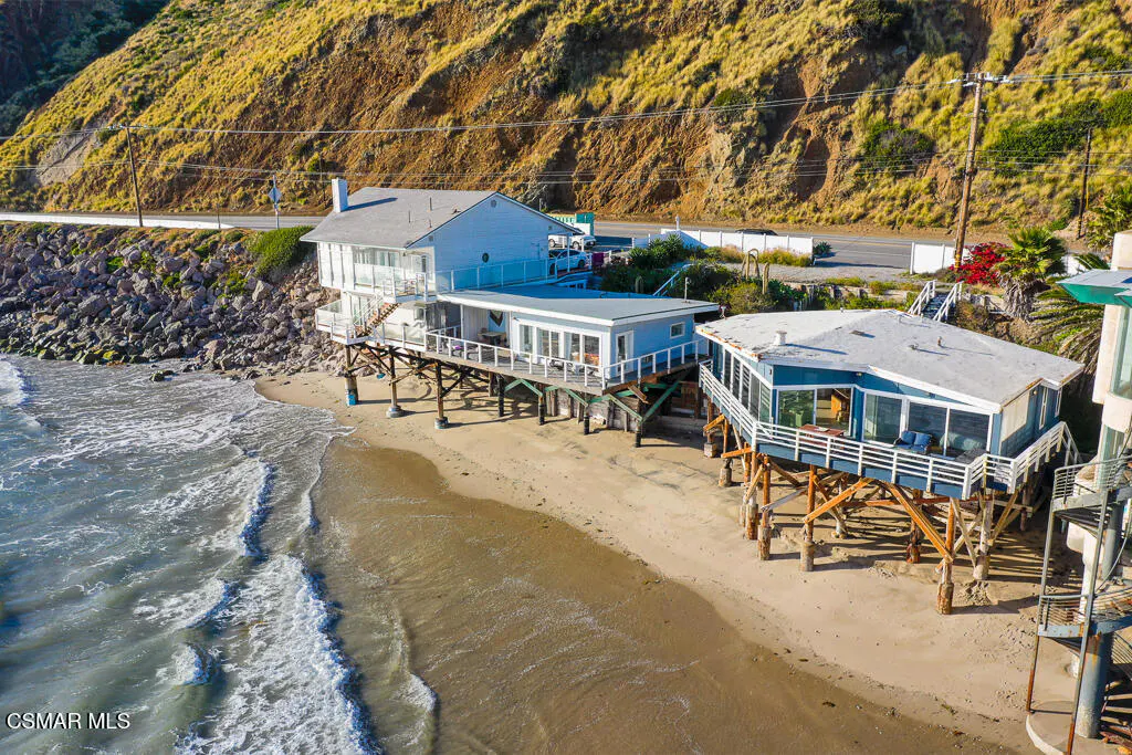 Aerial view of blue beach houses on stilts, with ocean waves, sandy beach, and a grassy hillside in the background.