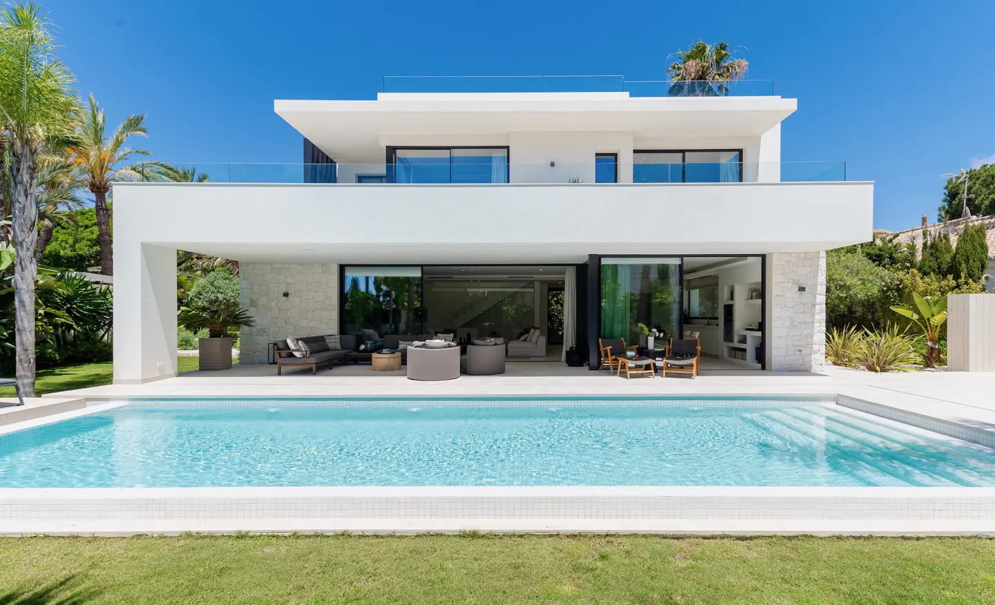 Two-story modern white house with a pool. Patio furniture is visible through sliding glass doors. Blue sky.