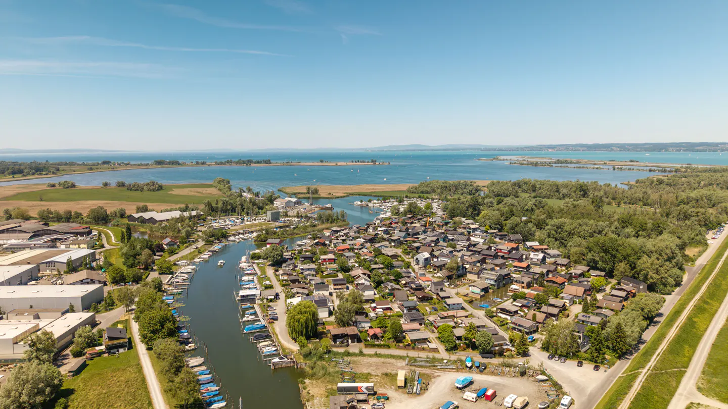 Aerial view of a lakeside community with small houses, a narrow canal filled with boats, and lush green trees under blue sky.