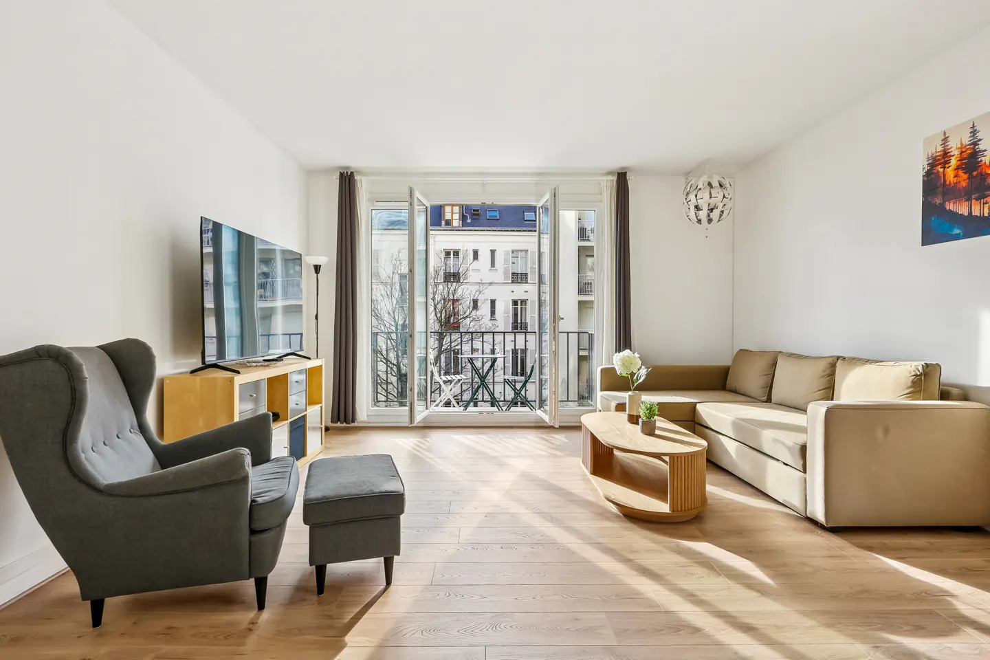 Bright living room with hardwood floors, gray armchair, beige sectional sofa, and open balcony doors.
