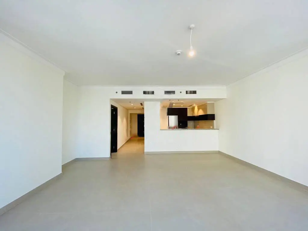 An empty, bright room with white walls and gray tile floors. An open kitchen area is visible in the background.