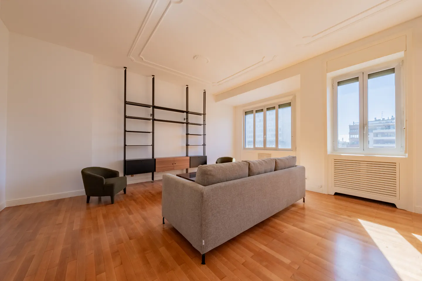 Bright living room with hardwood floors, white walls, and large windows. A gray sofa faces a black bookshelf. An olive chair sits to the left.