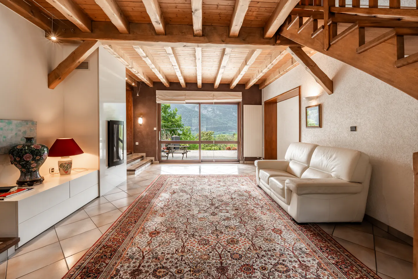 Living room with exposed wood beams, tile floor, and large rug. A white sofa sits near a sliding glass door with mountain views.