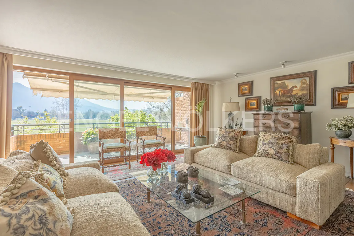 Bright living room with beige sofas, glass table, and balcony overlooking a green landscape. Art and furniture add character.