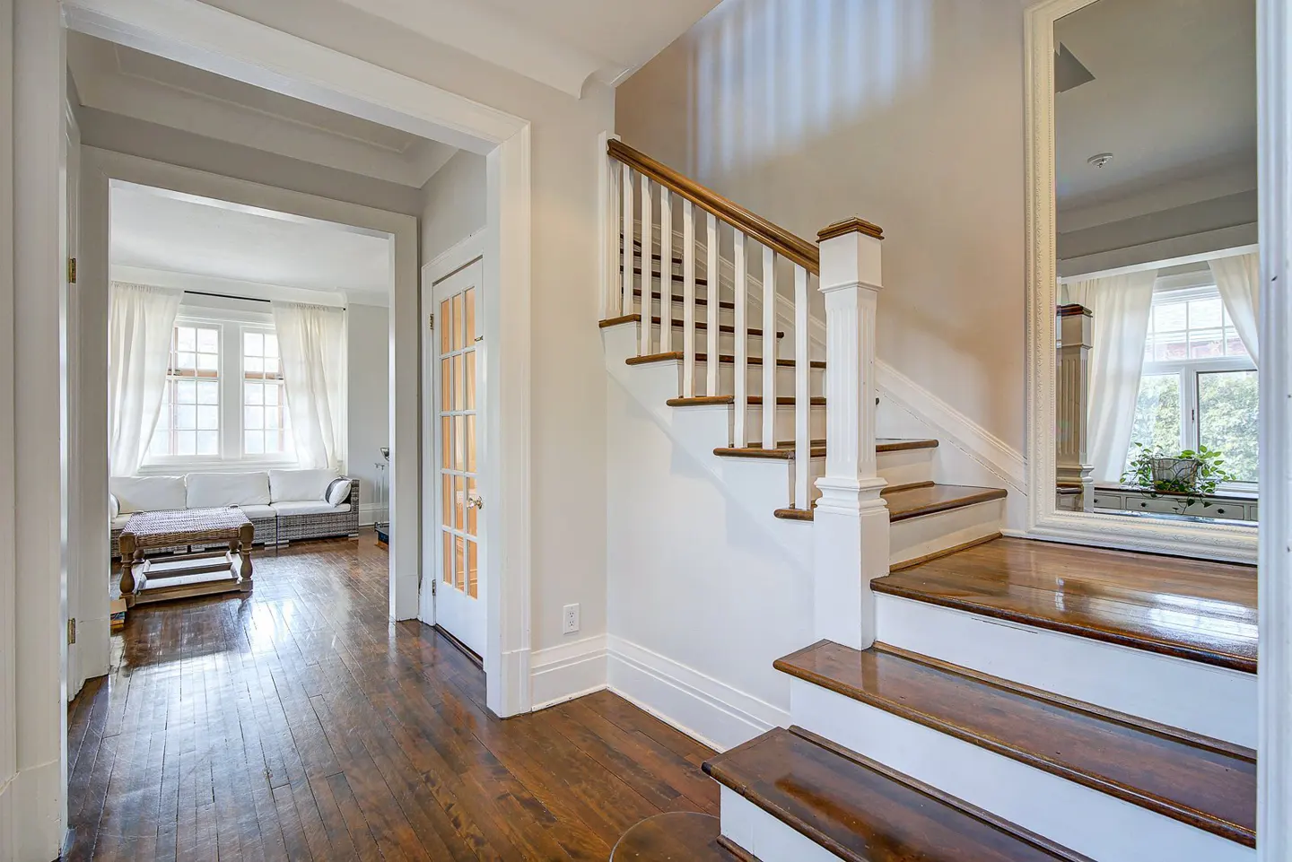 Bright foyer with hardwood floors, white walls, and a staircase with wood treads and white risers. A living room is visible through an archway.