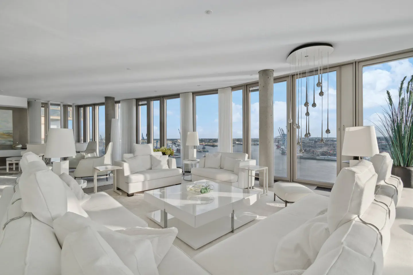Bright, modern living room with white sofas, chairs, and a glossy white table. Large windows offer a city and harbor view.