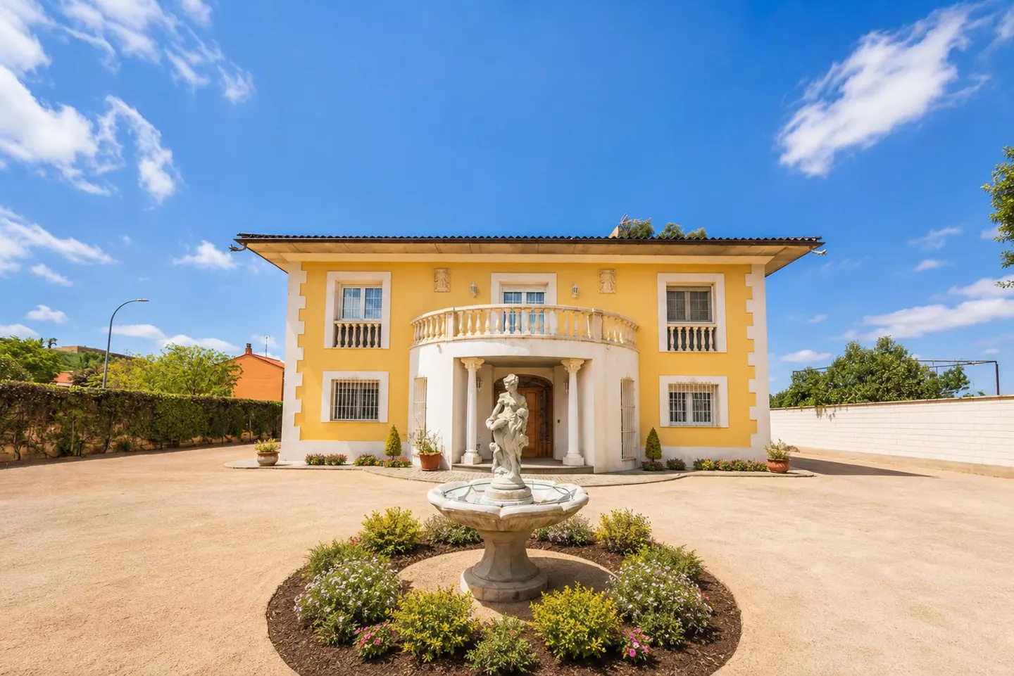Two-story yellow house with white trim, balcony, and a statue fountain in the front yard. Blue sky with clouds above.