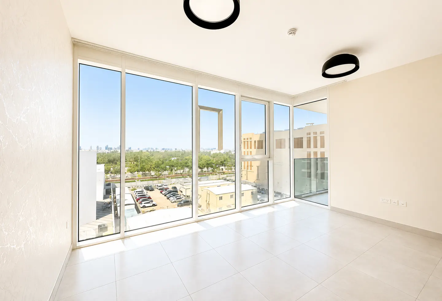 Bright, empty room with white tile floor and walls. Large windows offer a view of a parking lot, trees, and city skyline. Black, round light fixtures on the ceiling.