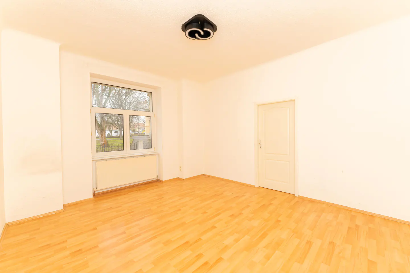 Bright, empty room with light wood floors, white walls, a window overlooking a yard, and a modern black ceiling light.