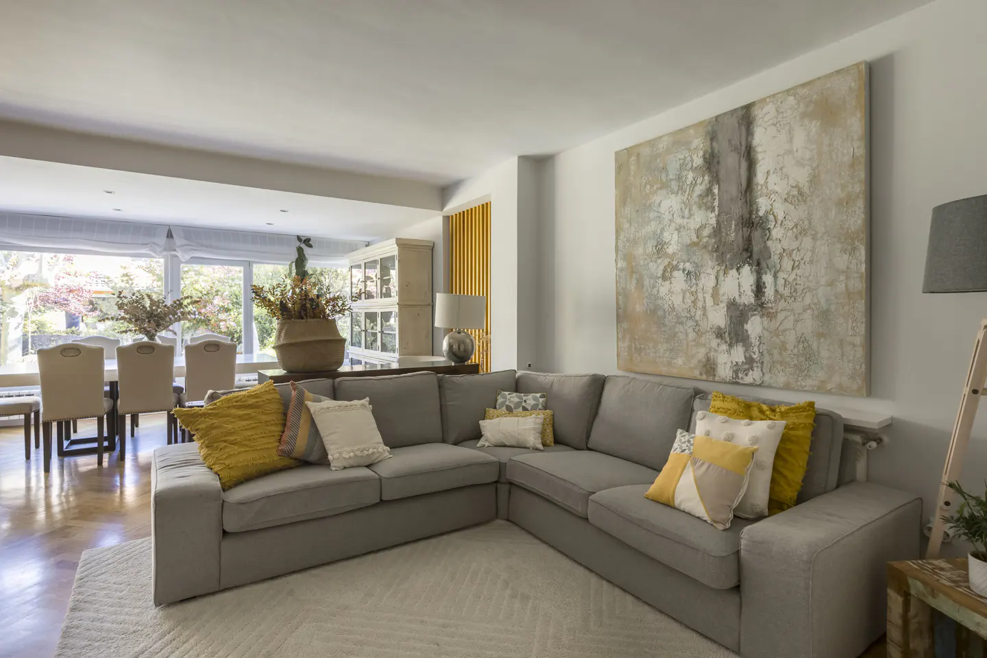 Living room with a gray sectional sofa, yellow pillows, and a large abstract painting on the wall. Dining table visible in the background.