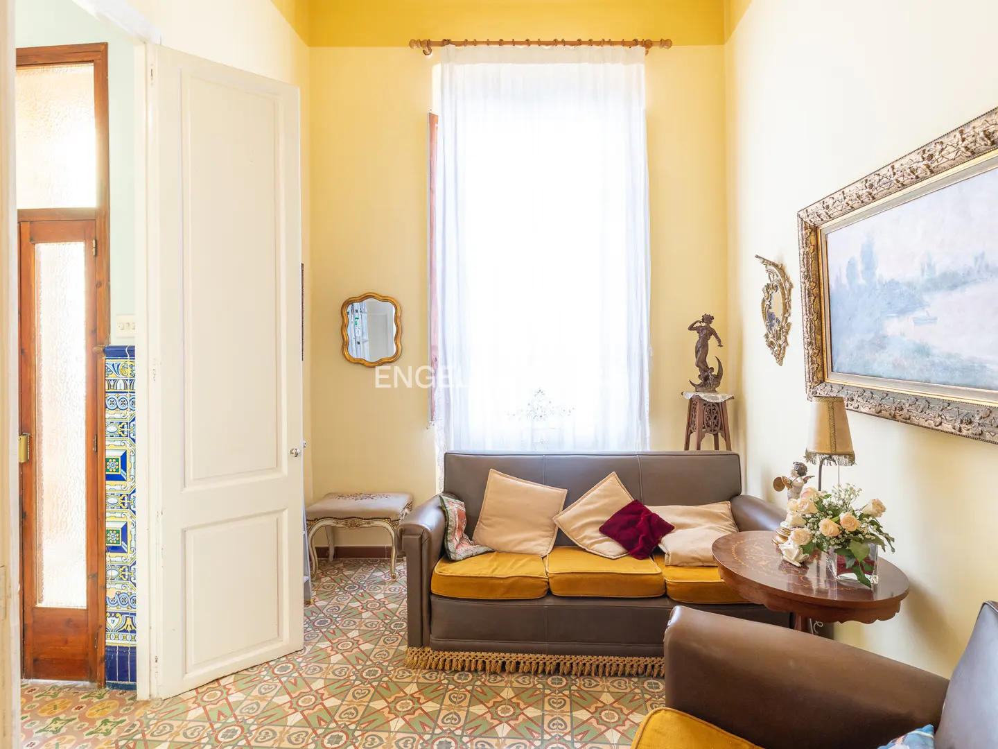 A living room with yellow walls, patterned tile floor, a brown sofa, and a round table with flowers.