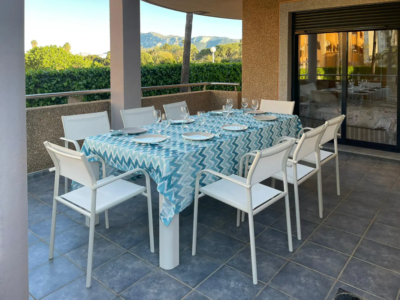 Outdoor patio with a dining table set for a meal. The table has a blue patterned tablecloth, plates, and glasses. White chairs surround the table.