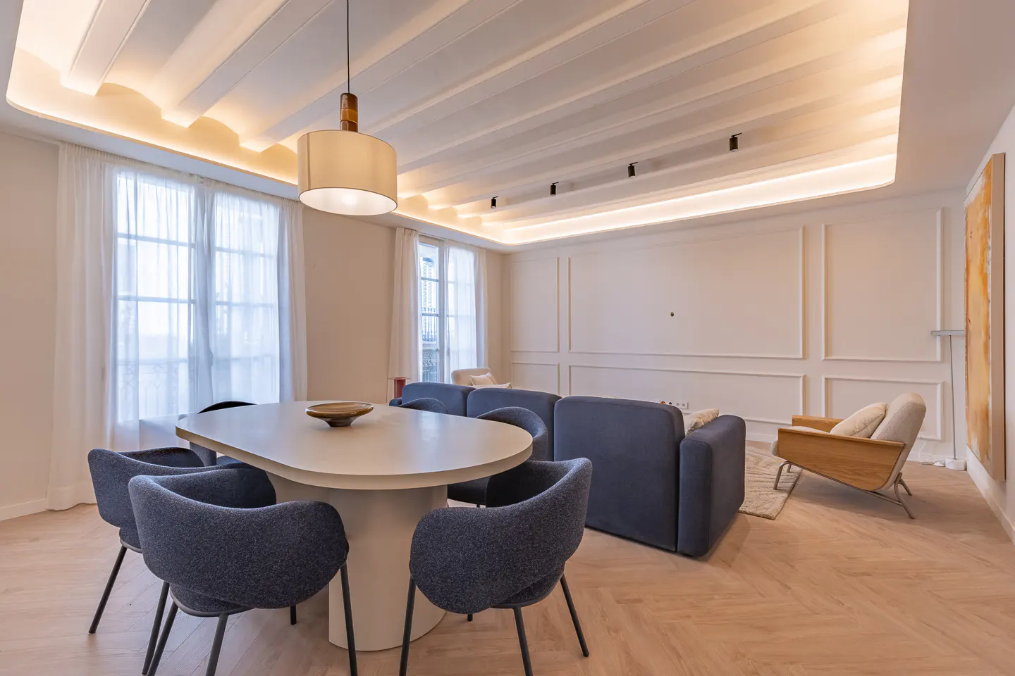 Bright, modern living room with a white table surrounded by blue chairs, a blue sofa, and a wooden chair. White walls and herringbone wood floors.