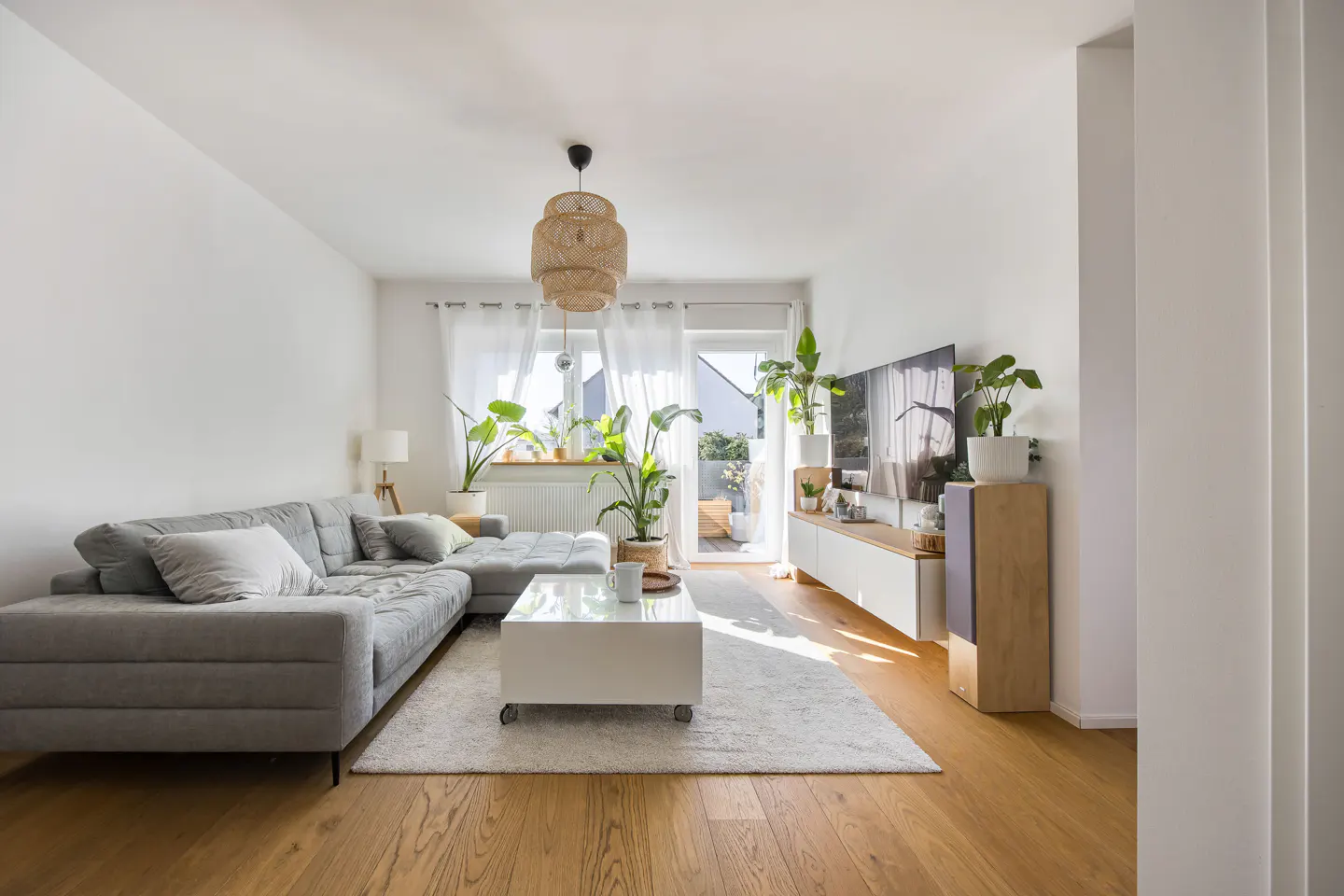 Bright living room with a gray sectional sofa, white coffee table, and a TV. Plants add greenery to the space. Hardwood floors and a woven light fixture complete the look.