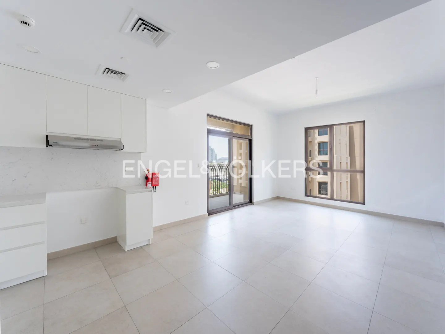 Bright, empty apartment with white walls, tile floors, and kitchen cabinets. A balcony and window offer city views.