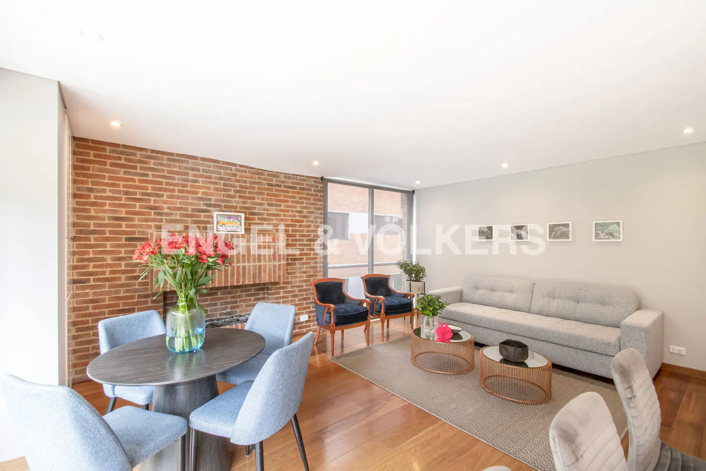 Living room with brick wall, gray sofa, blue chairs, and wood floors. A vase of red flowers sits on the dining table.