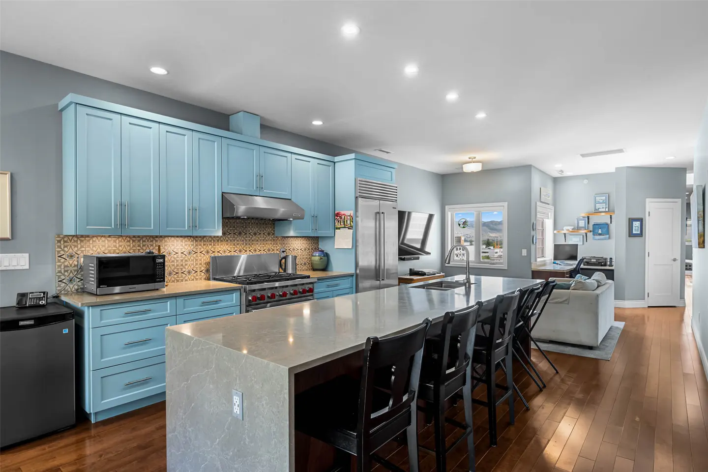 A modern kitchen with light blue cabinets, stainless steel appliances, and a large island with black stools. Hardwood floors lead to a living area.