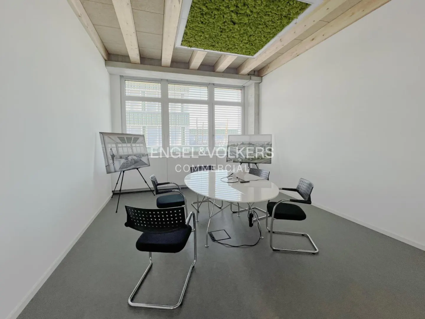 Bright office space with a white table, black chairs, and a green moss ceiling feature. Large windows provide natural light.