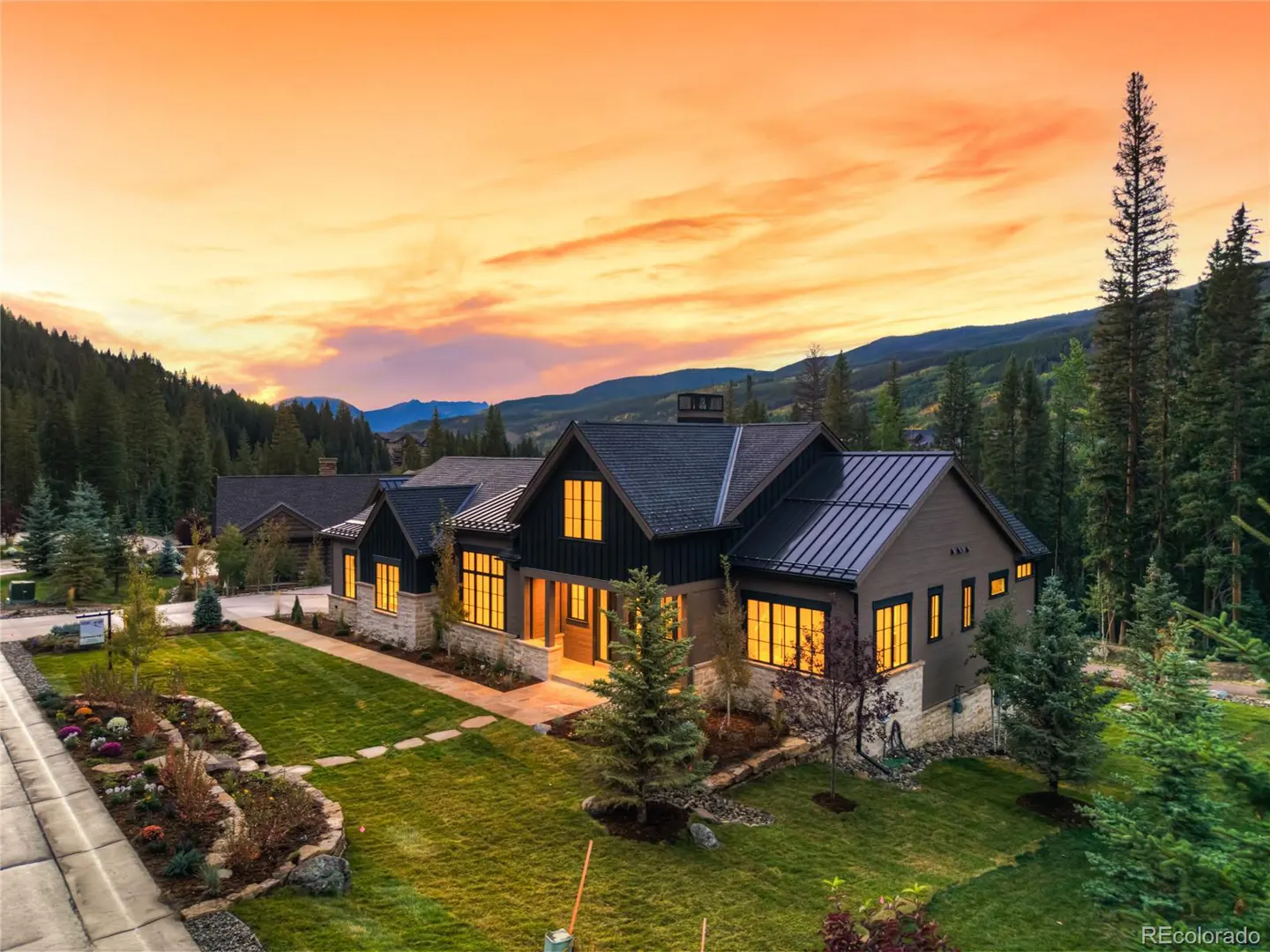 A two-story house with a dark roof and light-colored stone accents, set against a backdrop of mountains and a colorful sunset.
