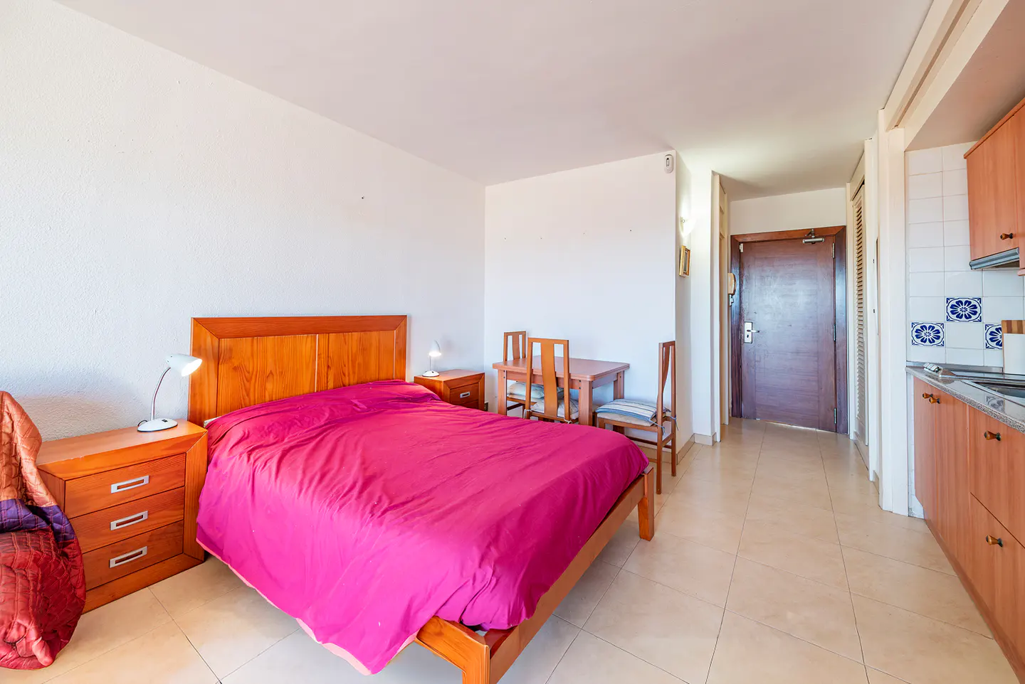 Studio apartment with a wooden bed covered in a pink sheet, a small dining table, and a kitchenette.