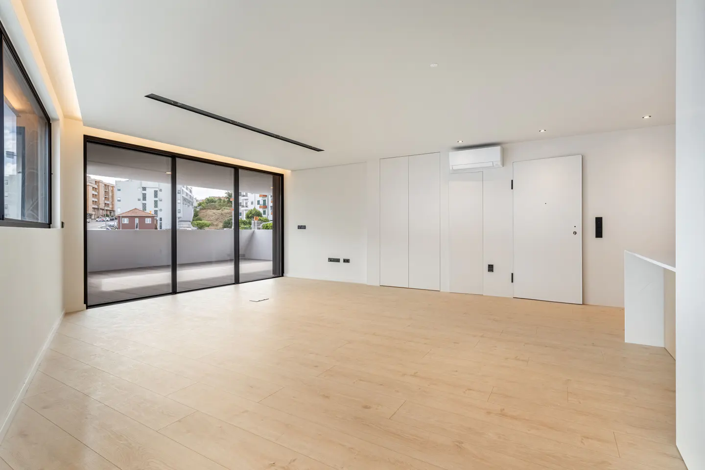 Minimalist empty room with light wood floors, white walls, and large sliding glass doors opening to a balcony.