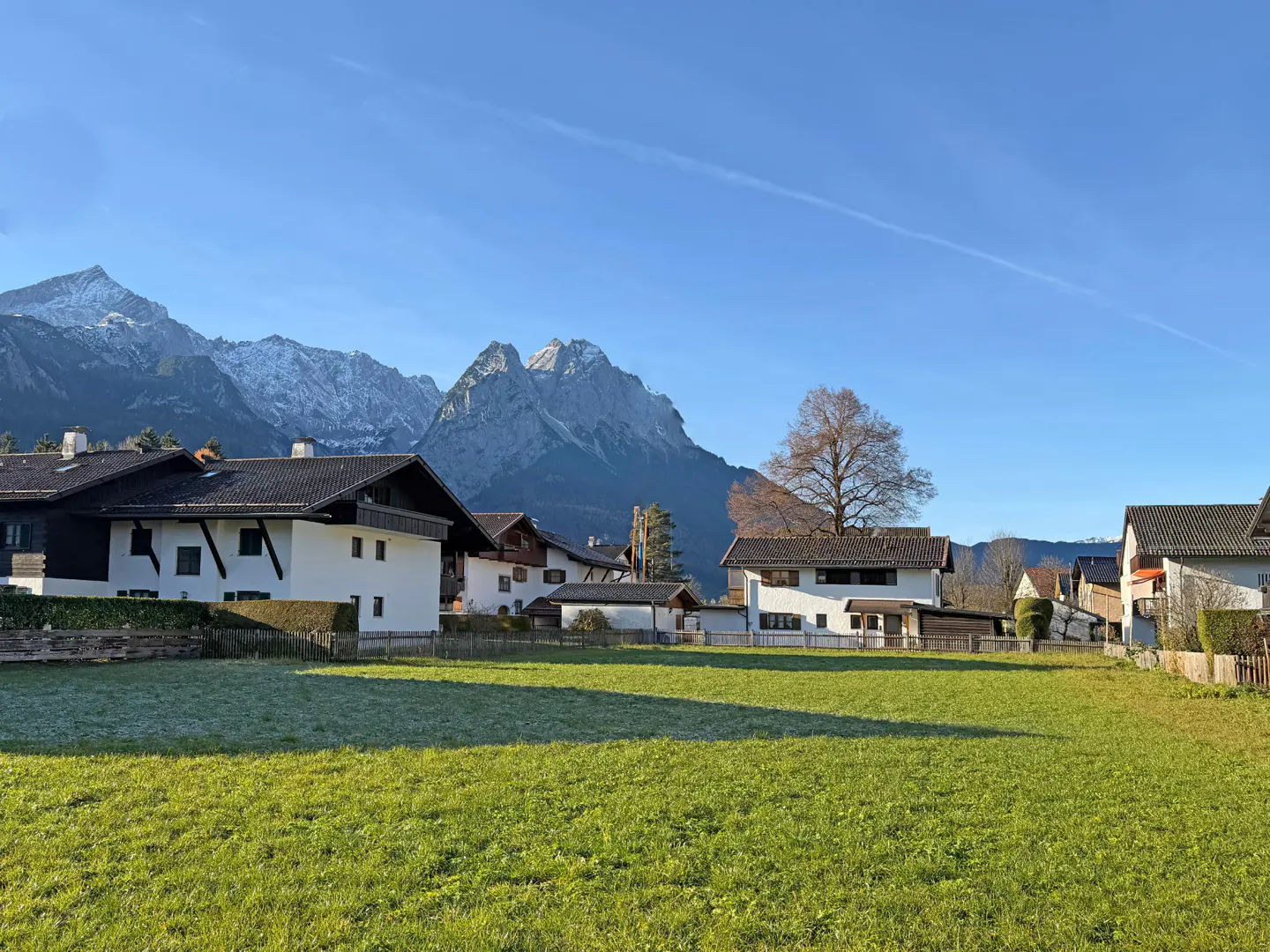 A scenic view of white houses with dark roofs, a green field, and snow-capped mountains under a blue sky.