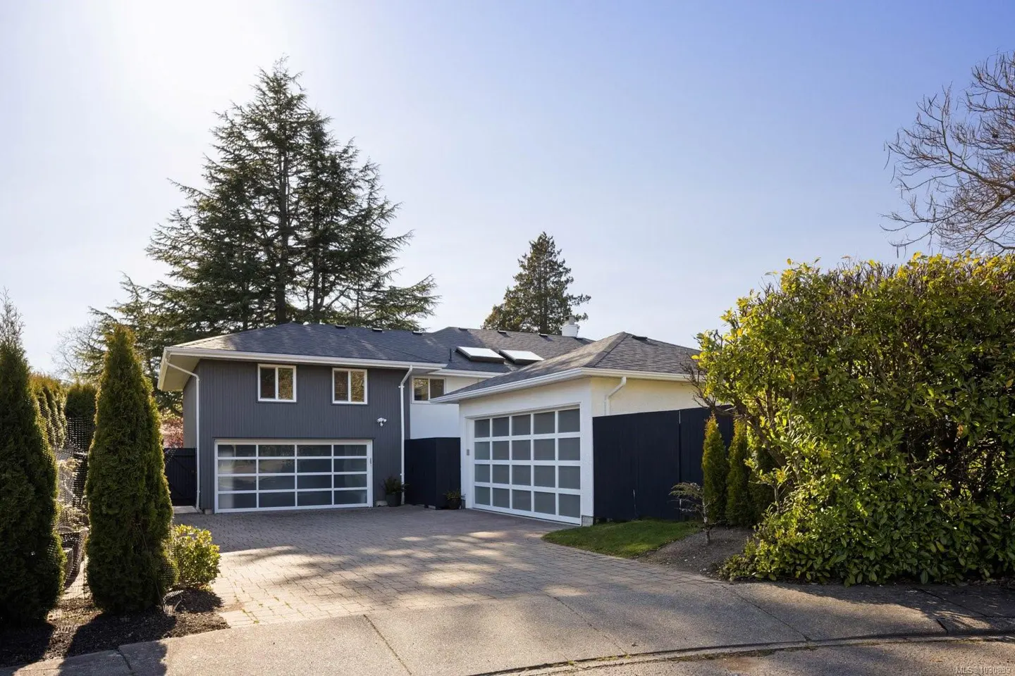 Exterior of a two-story gray house with white trim and two glass garage doors, a brick driveway, and green trees.