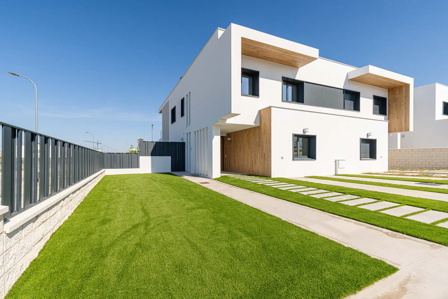 A modern two-story white house with a green lawn and a gray fence under a clear blue sky.