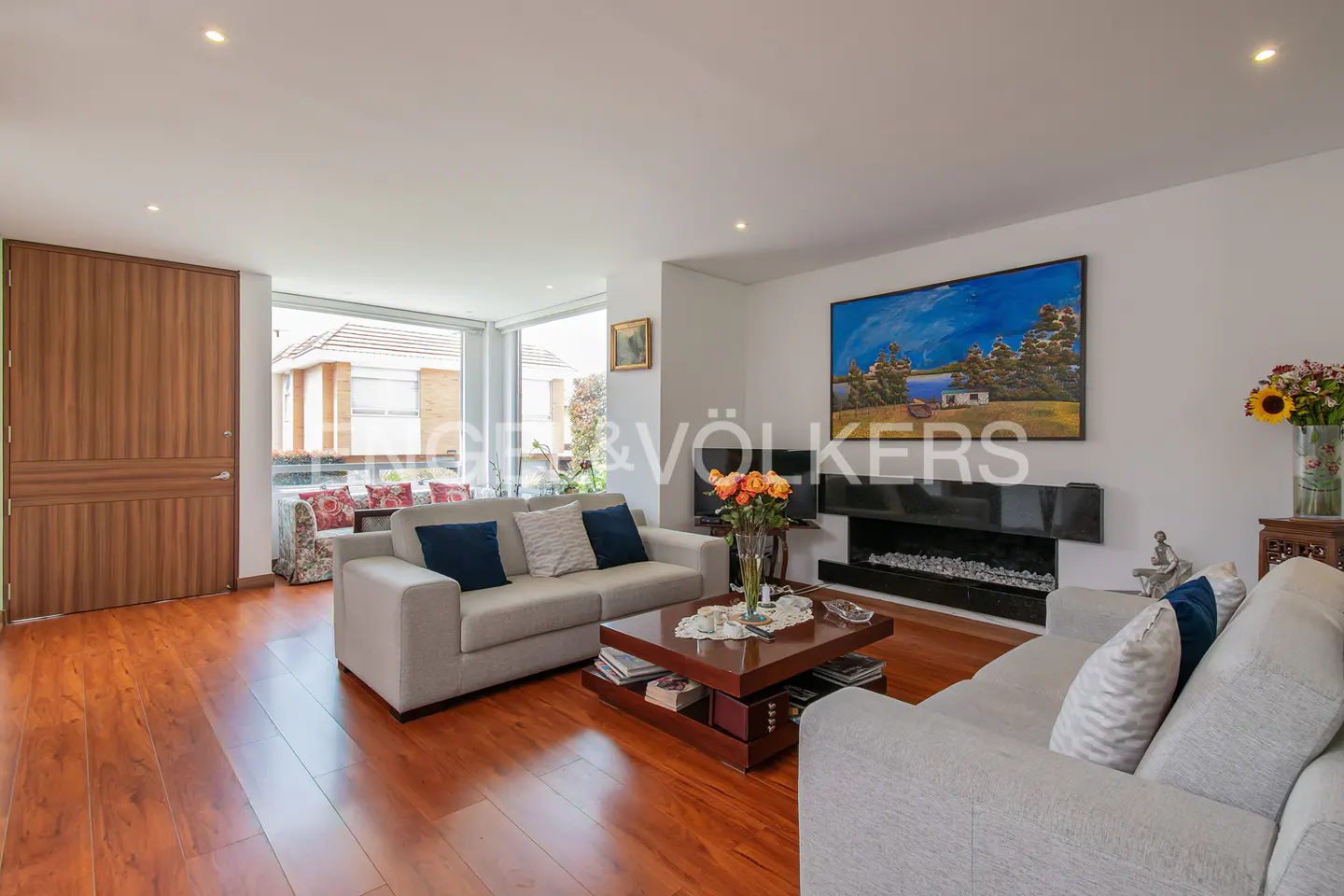 A bright living room with hardwood floors, two sofas, a fireplace, and a large painting. A wooden door is on the left.