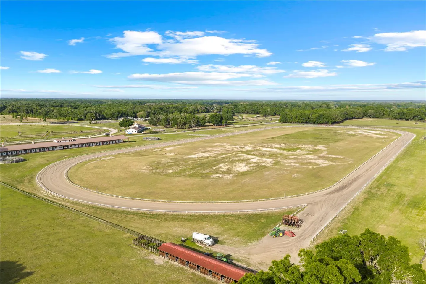 Aerial view of a horse farm with a dirt track, barns, green fields, and a blue sky with white clouds.