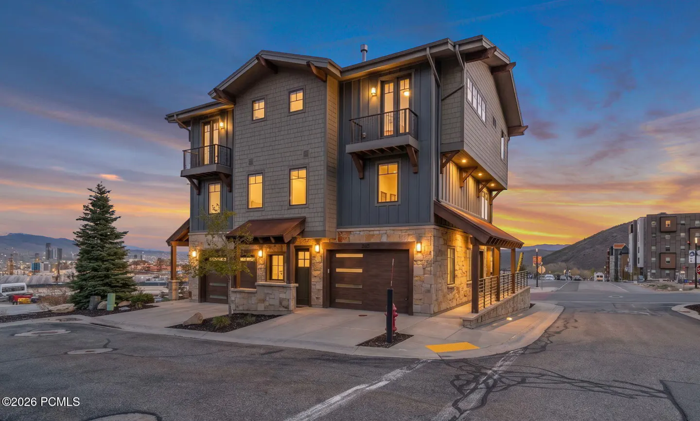 Three-story townhouse with gray siding and stone accents at dusk. Balconies, lit windows, and garage doors are visible.