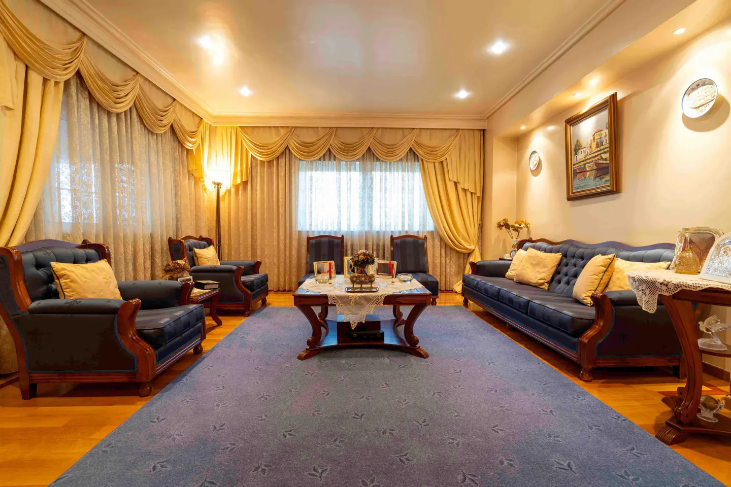 Living room with blue sofas, armchairs, and a patterned blue rug. Gold curtains frame the windows. A wooden coffee table sits in the center.