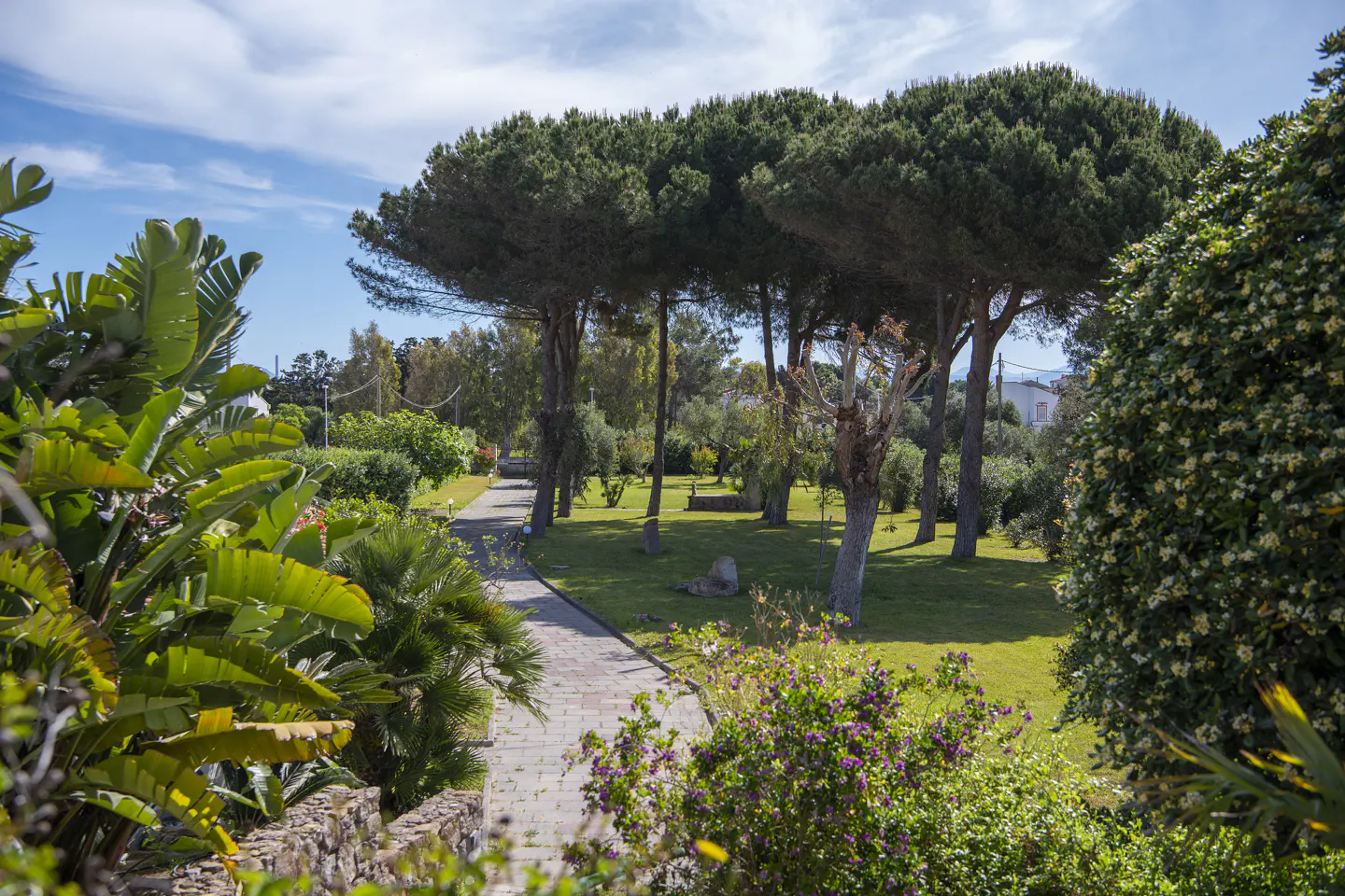 A brick path leads through a lush green garden with tall trees and flowering bushes under a blue sky.