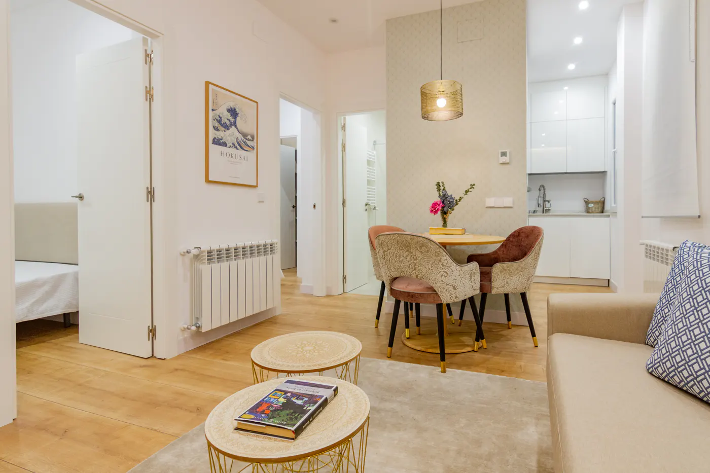 Bright apartment interior with wood floors, a round table with pink chairs, and a beige sofa with a blue patterned pillow. A Hokusai print hangs on the wall.