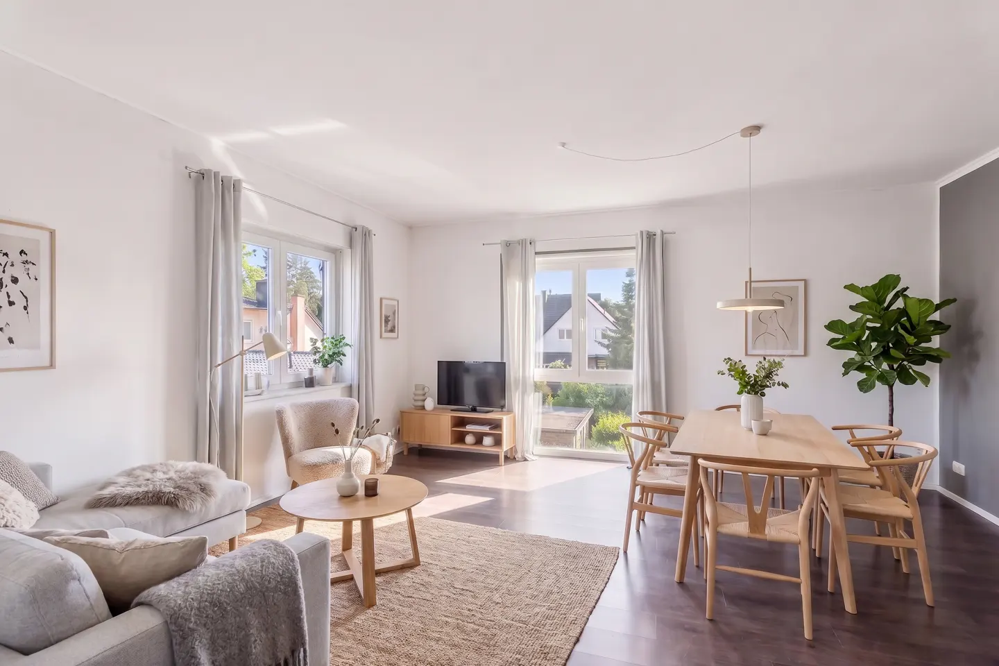Bright, open-plan living and dining area with white walls, wood floors, and large windows. A gray sofa, wood table, and chairs furnish the space.